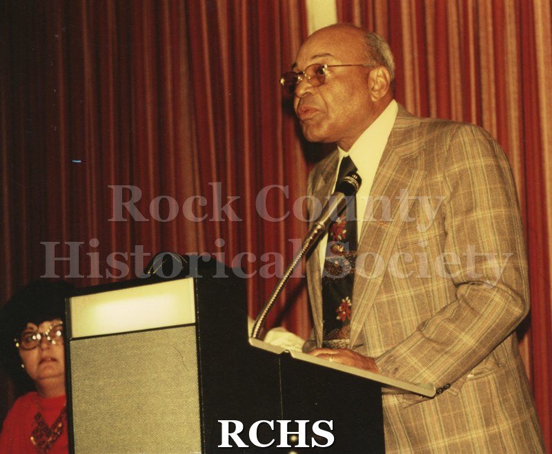 Colored photograph of John S. Scott standing in front of podium giving a speech. There is a woman seated on the left.