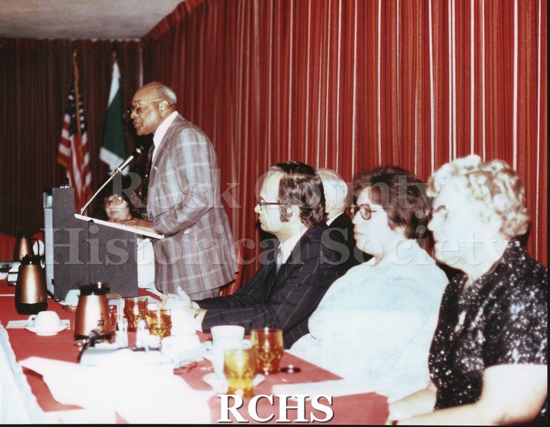 Colored photograph of John S. Scott standing in front of podium giving a speech. There are several people sitting to his right and another woman to his left.
