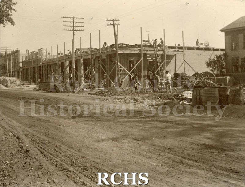 Black and white photograph of The Grange Store. Shows the construction of a building. There are men on the top working and men standing on the ground.