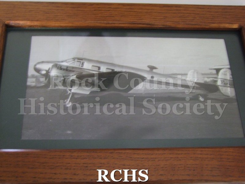 Black and white photograph of an airplane with twin propellers. Side view with "Parker" on propeller housing. Stripe along side of plane "Janesville/Wisconsin".