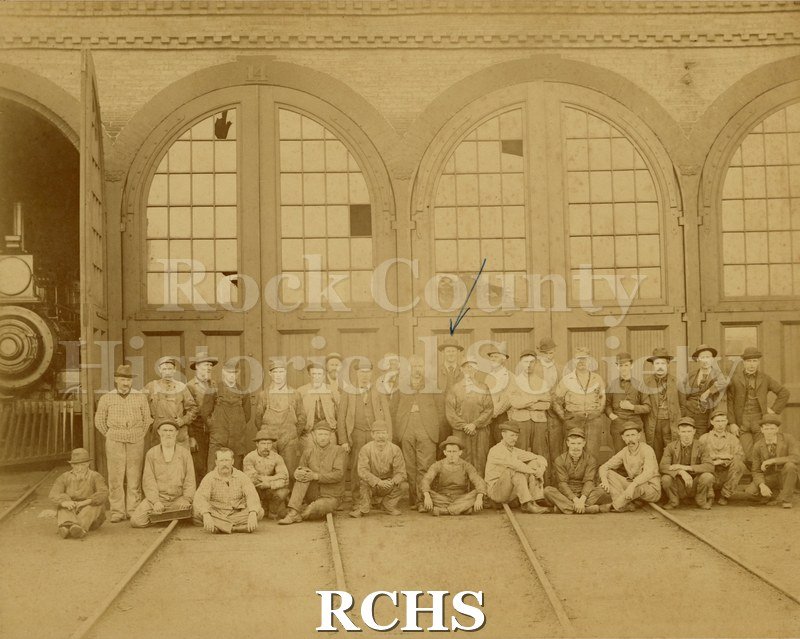 Black and white photograph of Old Engine House/Janesville WI. Shows railroad round house with 2 1/2 closed doors and 1/2 open door with engine inside.