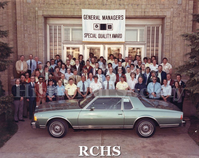 Color photo of employees standing in front of the Janesville General Motors plant beneath a banner that reads "General Manager's Special Quality Award." A large.