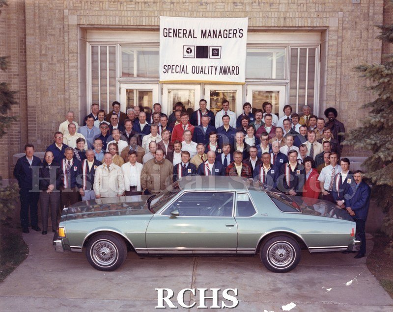 Color photo of employees standing in front of the Janesville General Motors plant beneath a banner that reads "General Manager's Special Quality Award." A large.