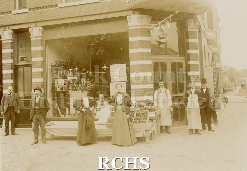 Black and white photograph of storefront with seven people standing in front of it. There is merchandise displayed in front of the store.