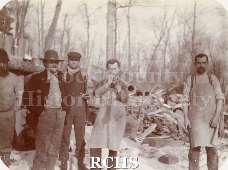 Black and white photograph of five unidentified men standing in front of a pile of logs.