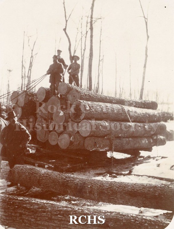 Black and white photograph of three unidentified men standing on top of a load of logs while another man stands in front of them.