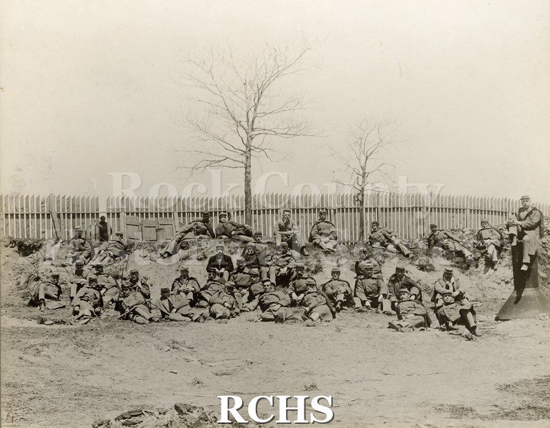 Black and white photograph of the Wisconsin National Guard. Soldiers are sitting and laying down in front of a fence.