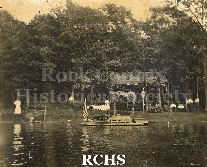 Black and white photograph of John Harlow & Otis Lincoln. Shows a cottage with a lake in front. There is a boat with a canopy over it moored at a pier.