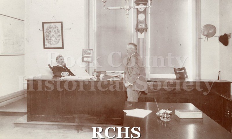 Photo. Black and white photograph of two unidentified men meeting in an office. Wall map of Beloit c. 1890-1900.