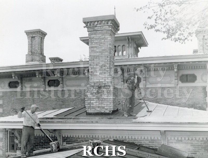 Black and white photograph of the Tallman House. Shows the chimney where two men are working on the roof area with large brushes.