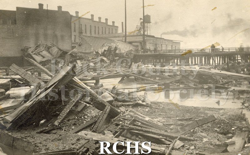 Postcard. Black and white photograph of Milwaukee St. Bridge Fire of 4/1/1913. Foreground of collapsed building, charred wood, large water puddles.