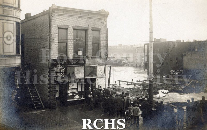 Postcard. Black and white photograph of 4/1/1913 Milwaukee St. Bridge. 2 standing buildings on left (one is "United Cigars"), people standing in road, 2 with bikes.