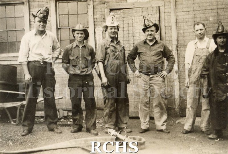 Black and white photograph of Milwaukee Street Bridge Fire 1942. Six men are standing in front of building. Five are wearing fireman helmets.