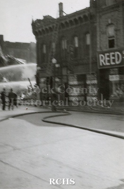 Black and white photograph of Milwaukee Street Bridge Fire 1942. Two standing buildings with two streams of water being sprayed to the left.