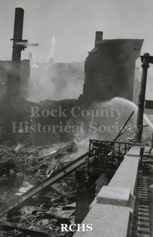 Black and white photograph of Milwaukee Street Bridge Fire 1942. Shows rubble and smoke, looking down from bridge. Standing buildings in background.