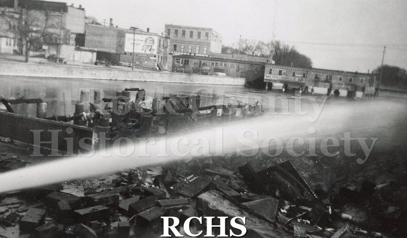 Black and white photograph of Milwaukee Street bridge fire 1942. Shows burned building debris with water stream from fire hose running diagonally across from left.