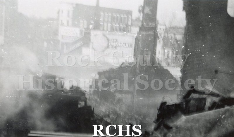 Black and white photograph of Milwaukee Street bridge fire 1913 showing burned building debris. Rock River and buildings visible in background.