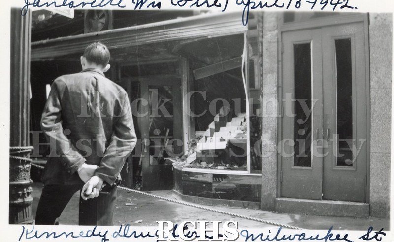 Black and white photograph of Janesville Fire 1942, Kennedy Drug Store showing open front of building with man standing with arms behind his back and street lamp.