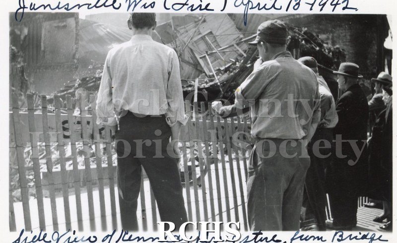 Black and white photograph of Milwaukee Street bridge fire 1942 showing collapsed building, fence in foreground with several men standing at fence.