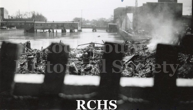 Black and white photograph of Milwaukee Street bridge fire 1942 showing smoke, river pilings and buildings in background.