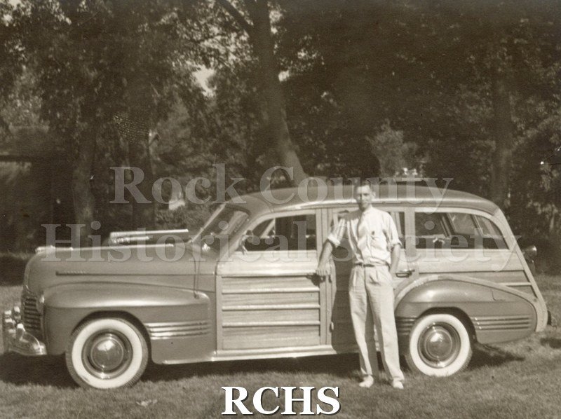 Black and white photograph of Robert Homberger in light colored clothes, standing beside a station wagon with his right hand on door handle and left hand behind.