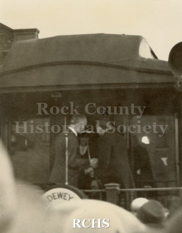 Black and white photograph of Thomas Dewey, one of two men in dark suits facing away from the camera on back of a train car.