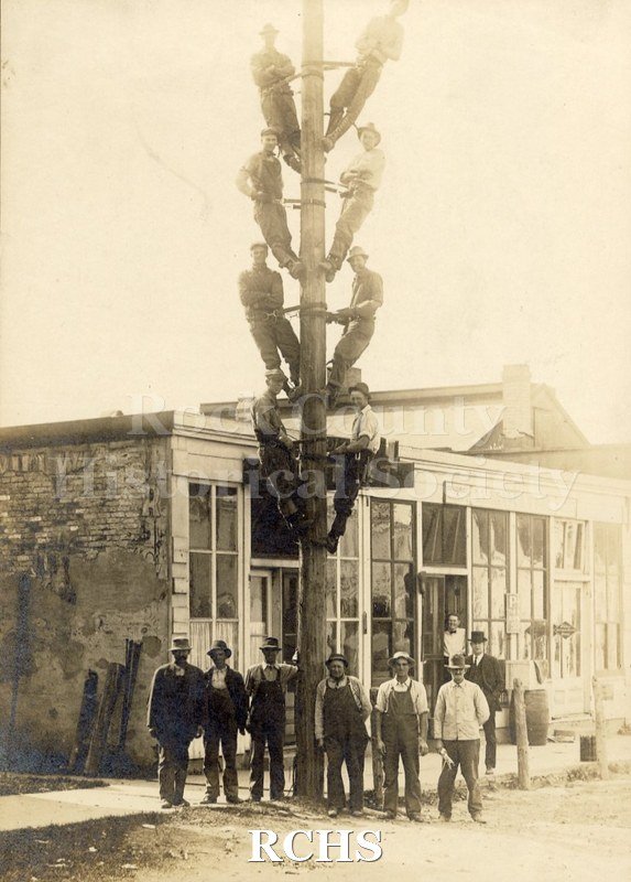 Black and white photograph of men of the Electrical Paver Crew, based in Clinton, 8 of whom are climbing on an electrical pole, the rest are standing on the ground.