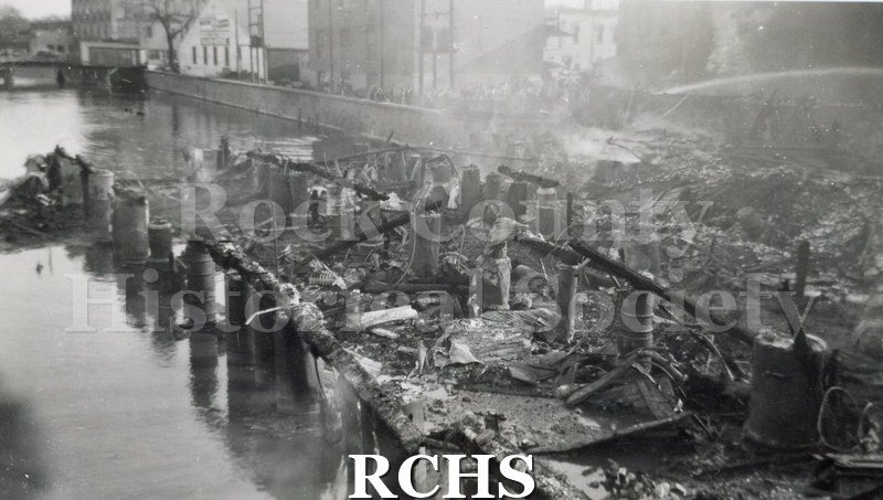 Black and white photograph of the Milwaukee Street Fire 4/13/42. It shows the charred remains of buildings and a fireman's hose streaming water.