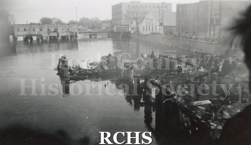 Black and white photograph of the Milwaukee Street Fire 4/13/42. It shows the charred remains of a building with pilings into the river.