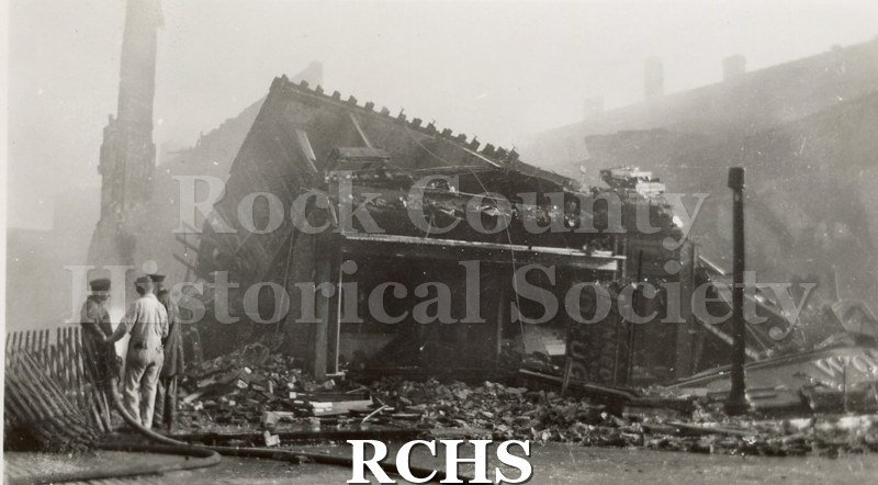 Black and white photograph of the Milwaukee Street Fire 4/13/42. It shows the charred remains of a building with three men standing on the left side with a hose.
