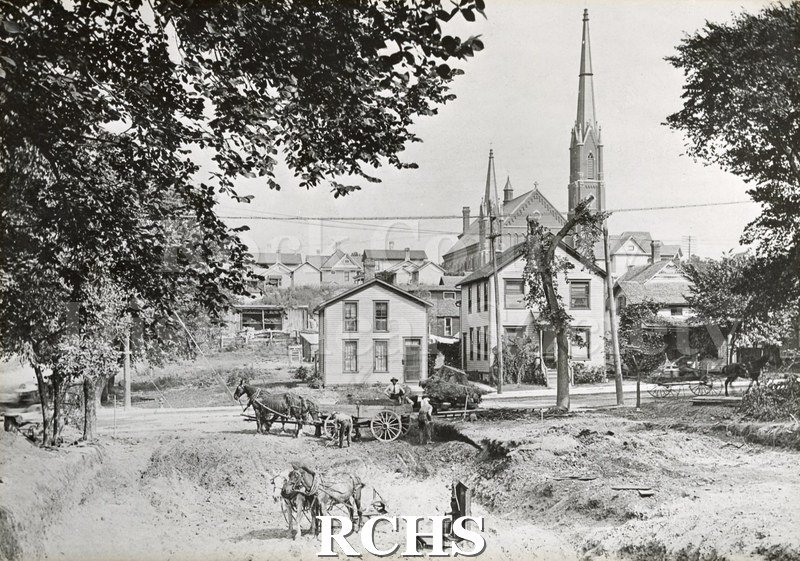 Black and white photograph of a construction site. Houses and a church steeple can be seen in the background.