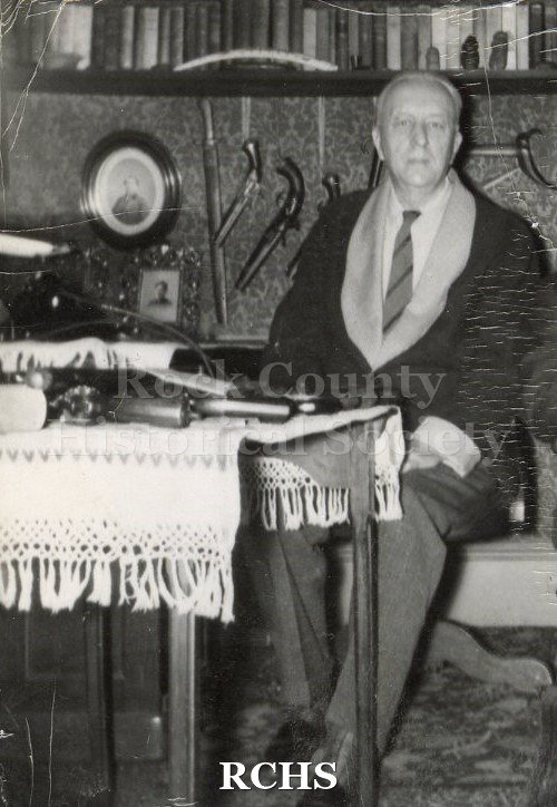 Black and white photograph of George K. Tallman sitting beside a table with a gun displayed. There are other items displayed behind him.