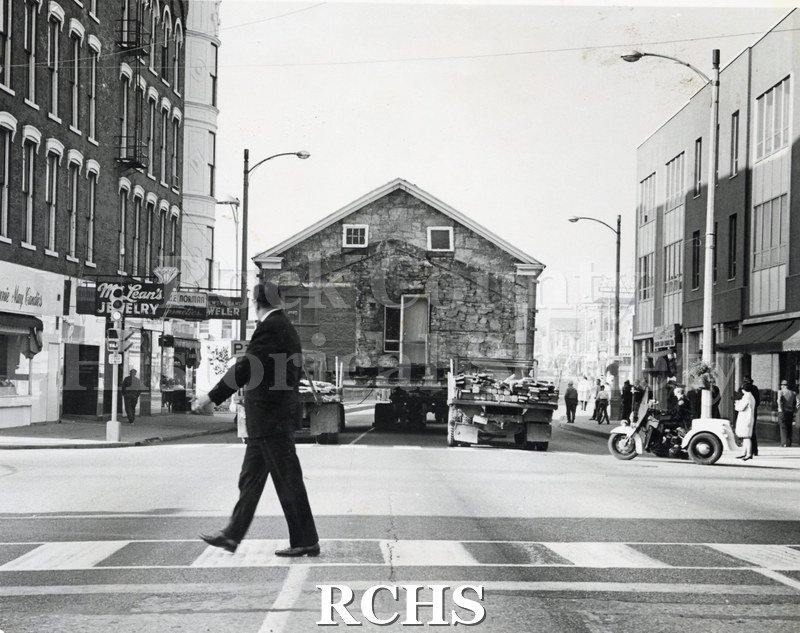 Black and white photograph of an unidentified man crossing the street looking at the Stone House on a trailer being moved along the street.