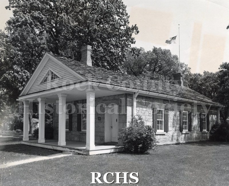 Black and white photograph of the Stone House with shrubbery and the American flag flying.