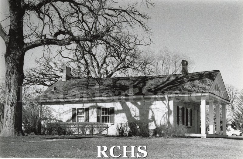 Black and white photograph of RCHS Stone House from the side with shrubbery and shadows of the trees on the side.