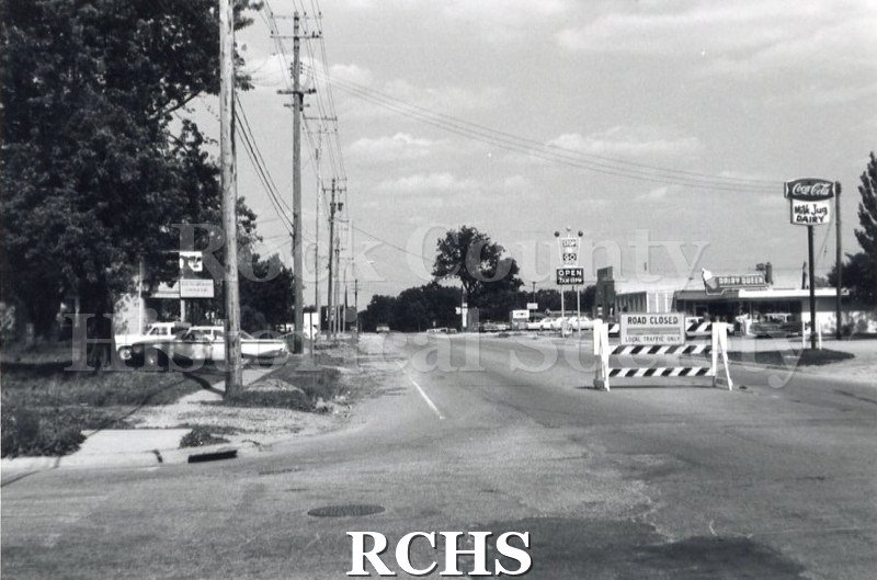 Black and white photograph of Racine Street with a sign: "Road Closed" in front of it. On the right is a sign for "Stop and Go" and another for "Milk Jug Dairy".