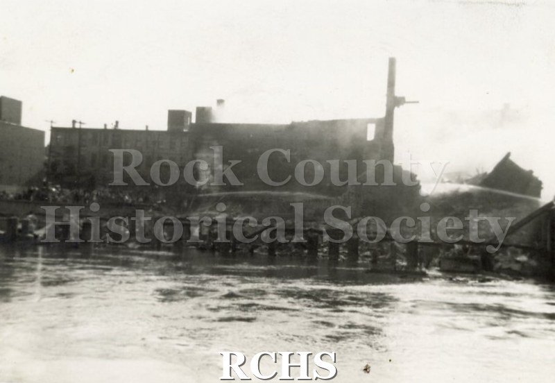 Black and white photograph of the Janesville Fire Milwaukee Street 1942. There is a large building with the river in front of it.