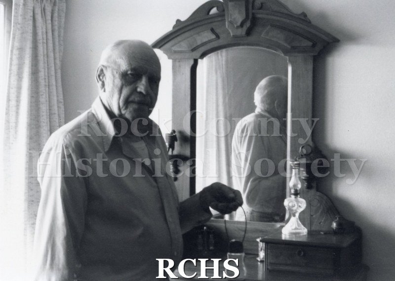 Black and white photograph of Art Levzow, Volunteer docent, standing by a dresser, holding a lamp. He is reflected in the mirror.