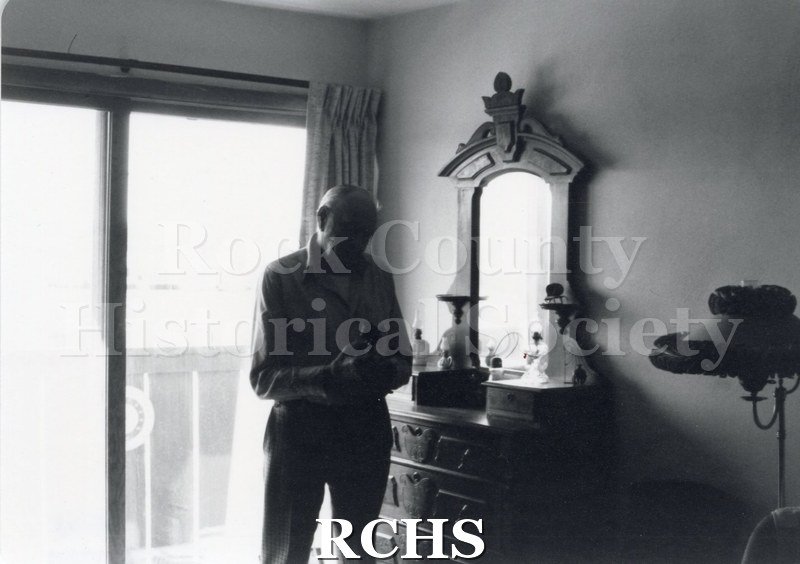 Black and white photograph of Art Levzow, Volunteer docent, standing by a dresser in the Tallman House.