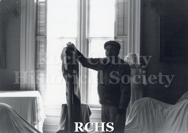 Black and white photograph of Art Levzow, volunteer docent, pointing to a statue in the Tallman House. Furniture is covered in white cloths.