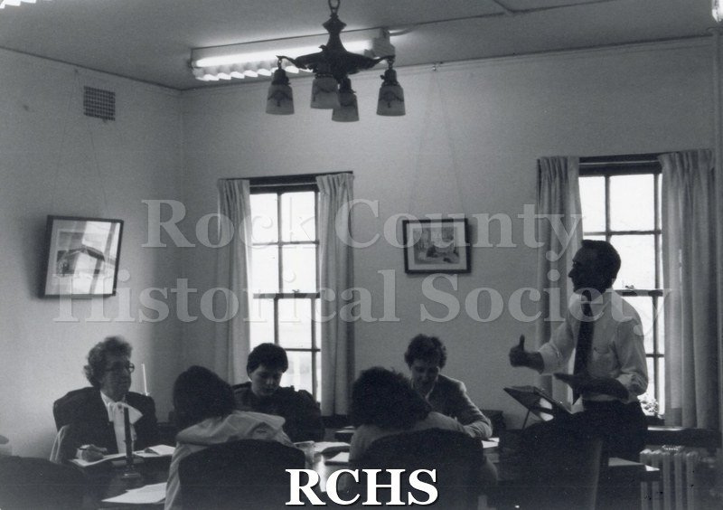Black and white photograph of Docent Training Session. Shows six people. There is a man standing at a lecturn talking to the people seated at the table.