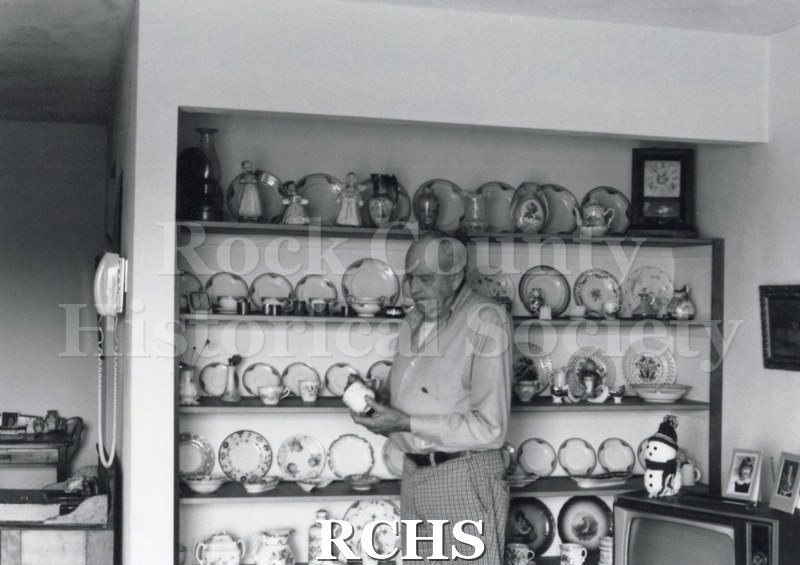 Black and white photograph of Art Levzow, volunteer docent, standing in front of a cabinet in which are displayed cups and saucers. He is holding a cup.