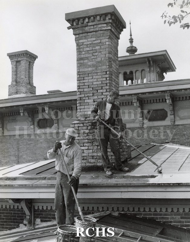 Black and white photograph of the roof of Tallman House. Two men are tarring the roofing in front of the chimney. Cupola is visible on top.