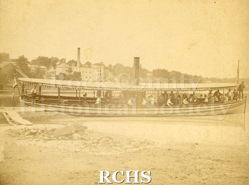 Sepia photo of steam launch "Columbia" with Buchholz boys. Two men stand at bow and stern. American flag displayed at bow. Boat is resting along shore.