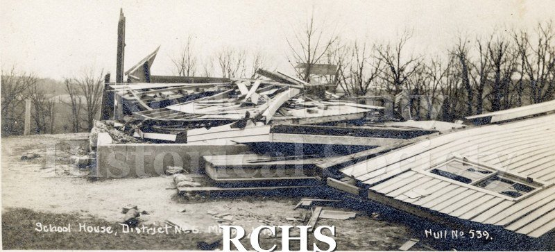 Postcard. Black and white photograph of 11/11/1911 cyclone. Collapsed building on foundation, broken windows, bare trees.