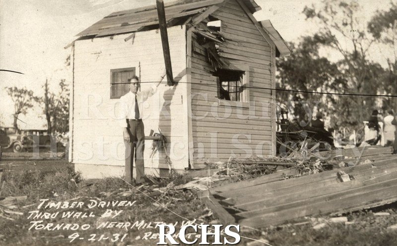 Postcard. Black and white photograph of 9/21/1931 tornado. Small white building with board sticking out side, man standing with hand on board.