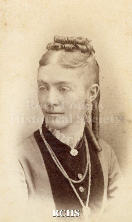 Black and white photograph of Kitty Van Allen. Above waist, head turned to her right. Hair center parted, loops on top near back, sausage curls from back.