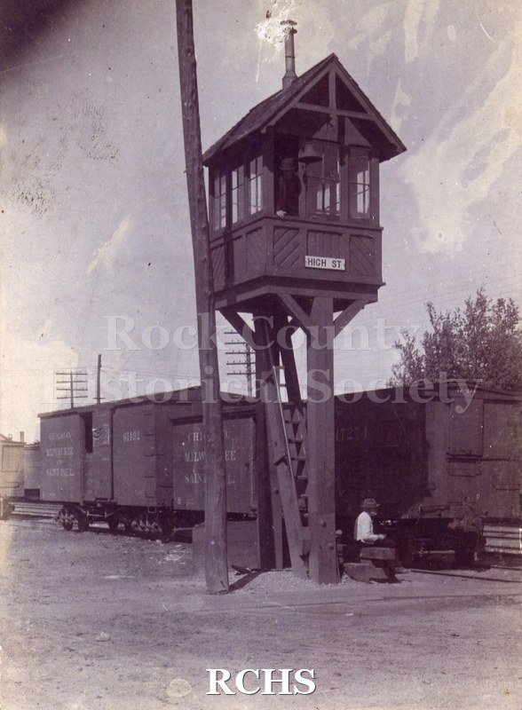 Black and white photograph of High Street Railroad Tower. Small building on posts with ladder underneath. Small chimney. Telephone pole to left.