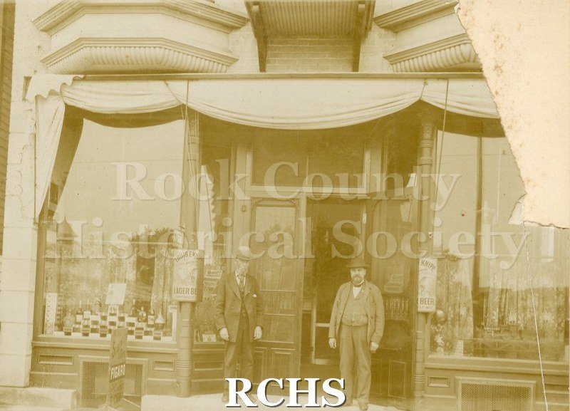 Black and white photograph of Smith's Hotel Bar. Shows two men standing in front of store front with one open screen door, one closed screen door and windows to.