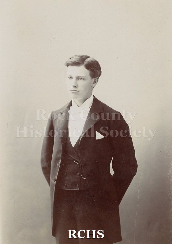 White cardstock cabinet card with scalloped edge. Cabinet card shows a Black and white photograph of a young man wearing a black suit and white shirt.